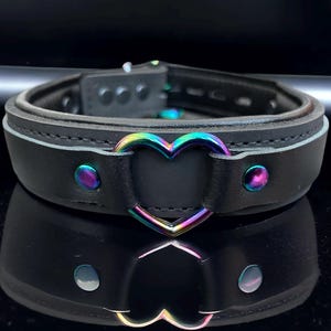May include: Black leather choker with a rainbow heart-shaped metal accent. The choker has a buckle closure and is decorated with silver studs.