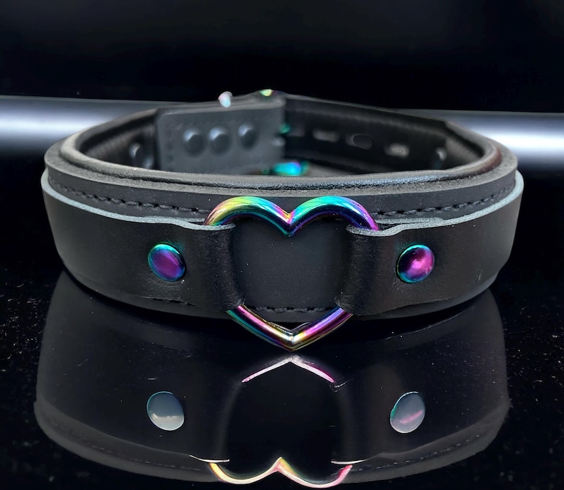 May include: Black leather choker with a rainbow heart-shaped metal accent. The choker has a buckle closure and is decorated with silver studs.