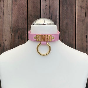 May include: Pink leather choker with a gold metal ring and the word "BIMBO" in gold lettering.