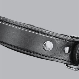 May include: Close-up of a black leather strap with silver-colored metal hardware. The strap is stitched along the edges and features a large, round silver rivet and a smaller rivet. The background is a solid gray.