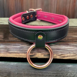 Puede incluir: Collar de cuero negro con forro rosa y un anillo de oro rosa. El collar tiene una hebilla y múltiples remaches.