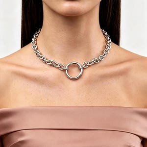 Könnte beinhalten: Eine silberfarbene Kettenhalskette mit einer Reihe gleichmäßiger ovaler Glieder, die mit einem zentralen, glatten kreisförmigen Ringanhänger verbunden sind. Die Kette liegt am Halsansatz und zeigt ein poliertes Metallfinish sowie einen Verschluss.