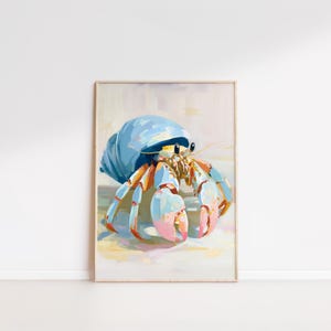 May include: A framed art print of a hermit crab. The crab has a light blue shell and orange and pink claws. The artwork is painted in a soft, impressionistic style, with a light background.