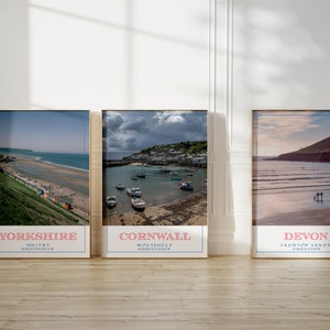 May include: Three framed prints of coastal scenes. The left print shows a beach with colorful beach huts and the text "Yorkshire Whitby". The middle print shows a harbor with boats and the text "Cornwall Mousehole". The right print shows a beach with people walking and the text "Devon Saunton Sands".