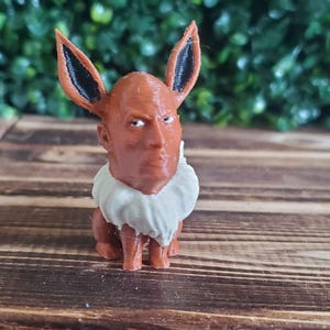 Op de afbeelding: Een 3D-geprint beeldje van een Pokémon-personage, Eevee, met het gezicht van Dwayne "The Rock" Johnson. Het beeldje is bruin en wit en staat op een houten oppervlak.