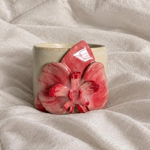 May include: A cylindrical ceramic cup with a smooth, off-white matte finish, featuring a large, three-dimensional sculpted orchid flower in shades of glossy pink and deep red attached to the front surface of the vessel.
