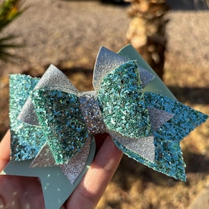 Pode incluir: Um laço de cabelo feito à mão com camadas de glitter verde-azulado e detalhes em glitter prateado. O laço é segurado em uma mão, mostrando seu design em camadas e textura brilhante. Um acessório de cabelo elegante.