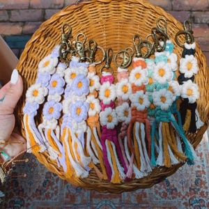 May include: A collection of macrame flower keychains featuring various colored petals including lavender, pink, orange, teal, and black, each with a white center and a brass-toned lobster clasp. The keychains have long, dangling fringe ends.