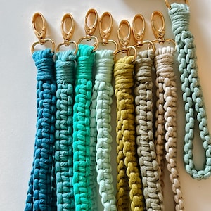 May include: A collection of eight macrame wristlet keychains in various colors including dark blue, teal, light green, mustard yellow, and beige. Each keychain features a gold-tone metal lobster clasp and is attached to a black car key fob.