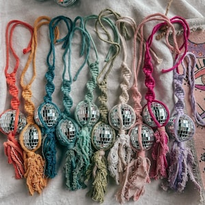 May include: A collection of nine hanging ornaments, each featuring a small mirrored disco ball encased in knotted cotton cord macrame. The cords come in various colors including orange, yellow, teal, green, cream, pink, and purple, with frayed tassels at the bottom.