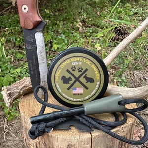 Könnte beinhalten: Ein Survival-Kit mit einem Messer mit braunem Griff, einem schwarzen Feuerstarter und einer Dose mit Kleintier-Überlebensschlingen. Die Dose ist schwarz mit weißem Text und Grafiken, einschließlich des Textes "GEN 2" und "MADE IN USA". Das Messer hat den Text "FIELDCRAFT" und "T-6830".