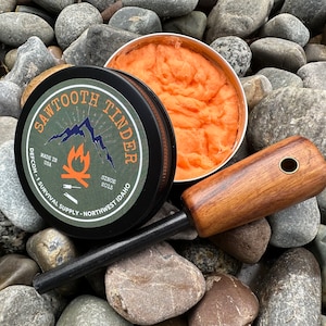 May include: A black tin with the text "Sawtooth Tinder" and "Made in USA" on it. The tin contains orange tinder. A black and brown fire starter is next to the tin.