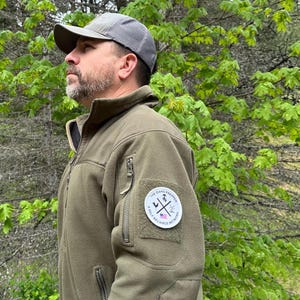 May include: An olive green fleece jacket with a gray cap. The jacket has a patch on the sleeve with the text "THE OHIO PREPPER" and a graphic. The jacket has a zipper closure and multiple pockets.