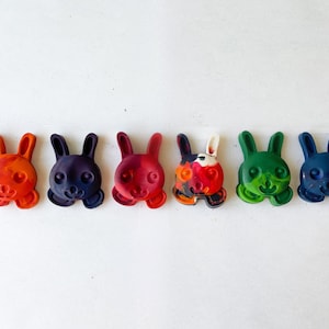 Pode incluir: Sete lápis de cera coloridos em forma de coelho em uma fileira. Os lápis de cera são vermelho, roxo, rosa, vermelho e branco, verde e azul.