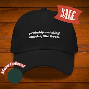 May include: Black baseball cap with white embroidered text that reads "probably watching Murder, She Wrote". The cap is on a wood background with a red "SALE" tag and a green circle with the text "More Colors!"