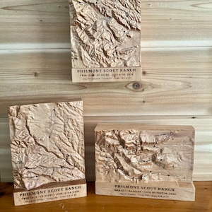 May include: Three light-colored wooden plaques featuring 3D topographical maps of Philmont Scout Ranch. Each plaque displays a carved mountain landscape with a black trail route, labeled with trek details, dates, troop numbers, and crew identifiers.