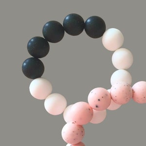 May include: A black, white and pink silicone bracelet with a speckled design. The bracelet is made of multiple beads and is in the shape of a figure of eight.