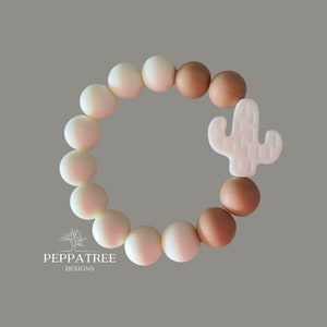 Puede incluir: Un brazalete de silicona blanco y marrón con un dije de cactus blanco. El brazalete está hecho de cuentas redondas y está diseñado para bebés que están dentiendo.