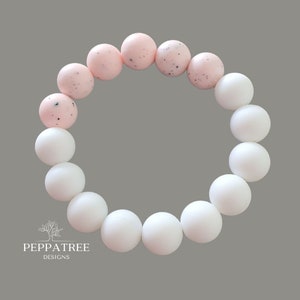 May include: A white and pink silicone bracelet with a speckled design. The bracelet is made of 12 beads, with 6 white beads and 6 pink beads.