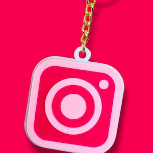 Op de afbeelding: Een roze en witte Instagram-logo sleutelhanger met een gouden ketting en ring.