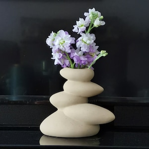 May include: A beige ceramic vase shaped like stacked rocks with a bouquet of purple and white flowers.