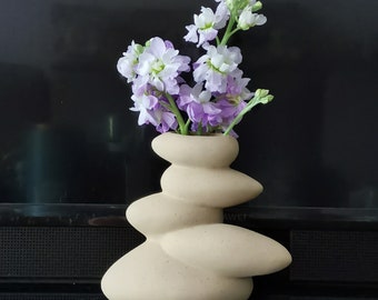 Op de afbeelding: Een beige keramische vaas in de vorm van gestapelde stenen met een boeket van paarse en witte bloemen.