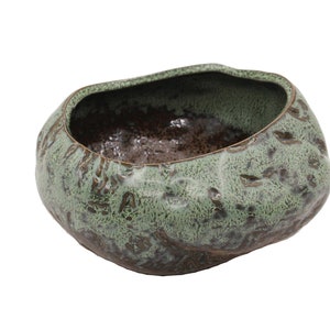 Op de afbeelding: Een handgemaakte keramische kom met een unieke, organische vorm. De buitenkant heeft een getextureerde, gevlekte groene glazuur, terwijl de binnenkant donkerbruin is. Deze decoratieve kom is ideaal voor woondecoratie.