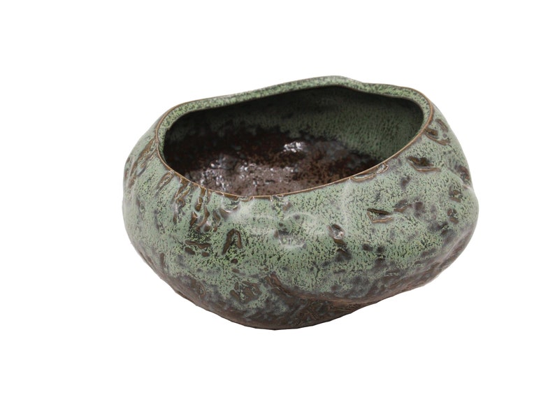 Op de afbeelding: Een handgemaakte keramische kom met een unieke, organische vorm. De buitenkant heeft een getextureerde, gevlekte groene glazuur, terwijl de binnenkant donkerbruin is. Deze decoratieve kom is ideaal voor woondecoratie.