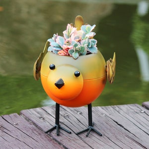 May include: A metal bird planter with a yellow and orange body, brown wings, and black legs. The planter is filled with green and pink succulents.
