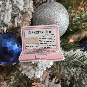 May include: A pink laptop sticker with the word "Dissertation" on the screen, decorated with black lines and text, is displayed on a Christmas tree. The background includes blue and silver ornaments and snow-covered branches.