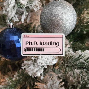 May include: A pink sticker with the text "Ph.D. loading" in a computer window design. The sticker is placed on a Christmas tree with blue and silver ornaments and frosted branches.