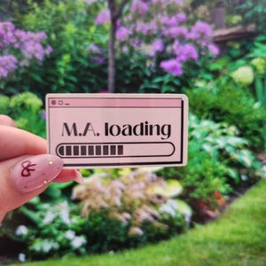 Può includere: Un adesivo rettangolare con sfondo rosa e bordo nero. L'adesivo recita "M.A. loading" in testo nero, con un grafico della barra di caricamento sottostante. L'adesivo è tenuto davanti a uno sfondo sfocato del giardino.