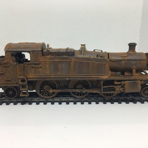 May include: A rusty model train engine on a track. The engine is brown and has a black chimney. The engine has six wheels and is made of metal.