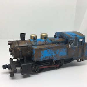 Puede incluir: Una locomotora de tren de juguete azul y marrón con una ventana rota. La locomotora tiene una chimenea negra y detalles dorados.