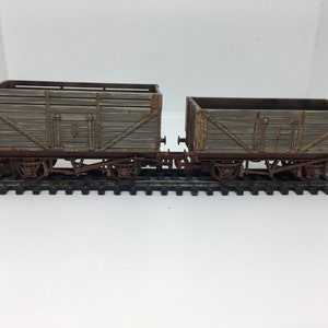 May include: Two wooden boxcars on a model train track. The boxcars are weathered and have rusty metal accents. The boxcars are connected together and are on a black track.