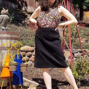 May include: A woman wearing a black skirt and a floral print top stands in a garden setting. She is smiling and looking at the camera. There is a wooden barrel, a windmill, and a small owl statue in the background.