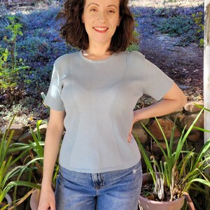 May include: A woman wearing a light blue ribbed knit top with short sleeves and blue jeans. She is standing in front of a green leafy plant and a wooden fence.