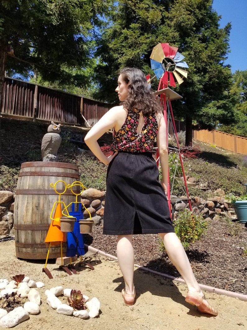 May include: A woman wearing a floral print tank top and a black skirt stands in a garden setting. She is looking to the left of the frame. There is a wooden fence, a barrel, and a metal windmill in the background.