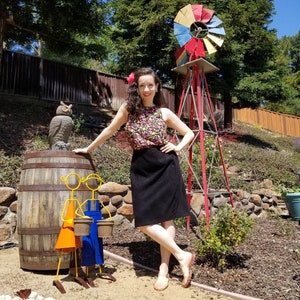 May include: A woman in a floral print top and black skirt stands in a garden setting. She is leaning against a wooden barrel with two metal figures holding buckets. A red windmill with colorful blades is in the background.