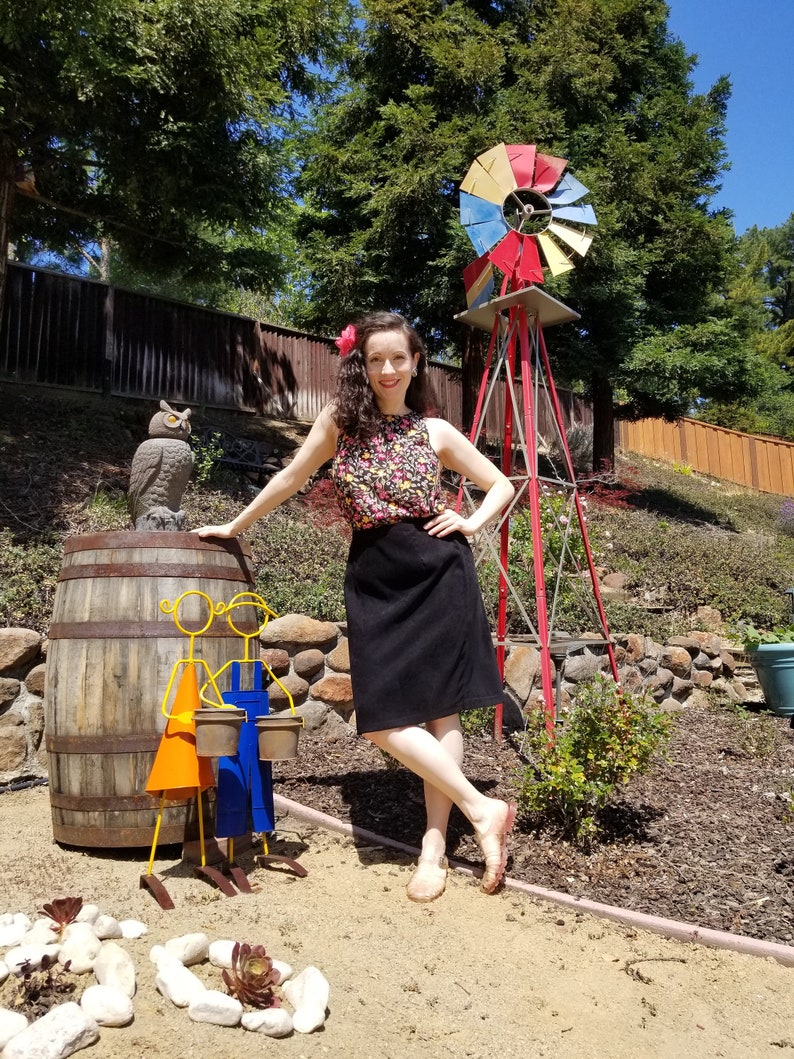 May include: A woman in a floral print top and black skirt stands in a garden setting. She is leaning against a wooden barrel with two metal figures holding buckets. A red windmill with colorful blades is in the background.