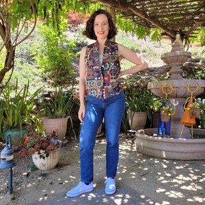 May include: A woman wearing a multi-colored patterned vest with a blue denim shirt underneath, blue jeans, and white sneakers. She is standing in a garden setting with a stone fountain in the background.