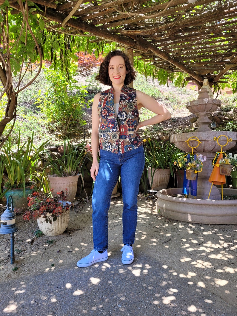 May include: A woman wearing a multi-colored patterned vest with a blue denim shirt underneath, blue jeans, and white sneakers. She is standing in a garden setting with a stone fountain in the background.