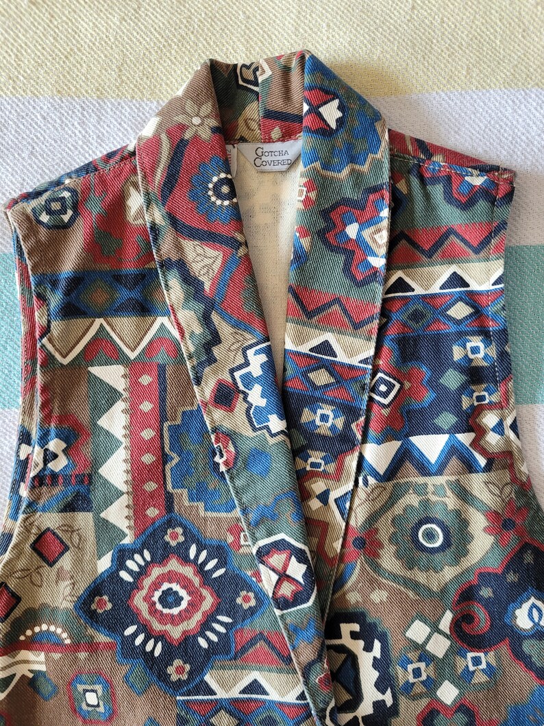 May include: A sleeveless vest with a geometric pattern in shades of brown, red, blue, and white. The vest has a large collar and a label that reads "Gotcha Covered".