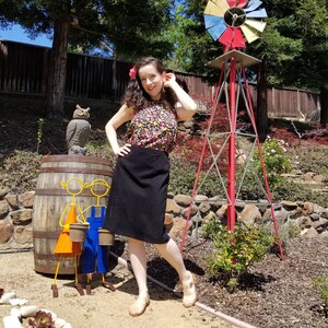 May include: A woman wearing a floral print top and a black skirt stands in a garden setting. She is smiling and looking at the camera. Behind her is a red windmill with a yellow, blue, and red design. There is a wooden barrel with a metal sculpture of two children holding buckets in front of her.