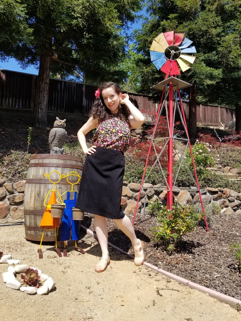 May include: A woman wearing a floral print top and a black skirt stands in a garden setting. She is smiling and looking at the camera. Behind her is a red windmill with a yellow, blue, and red design. There is a wooden barrel with a metal sculpture of two children holding buckets in front of her.