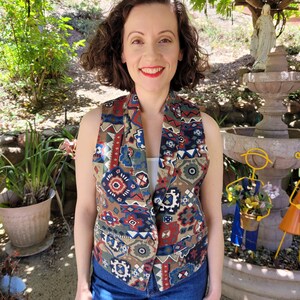 May include: A sleeveless vest with a colorful geometric pattern in shades of red, blue, green, and brown. The vest has a button closure and two pockets.