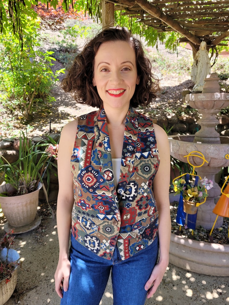 May include: A sleeveless vest with a colorful geometric pattern in shades of red, blue, green, and brown. The vest has a button closure and two pockets.