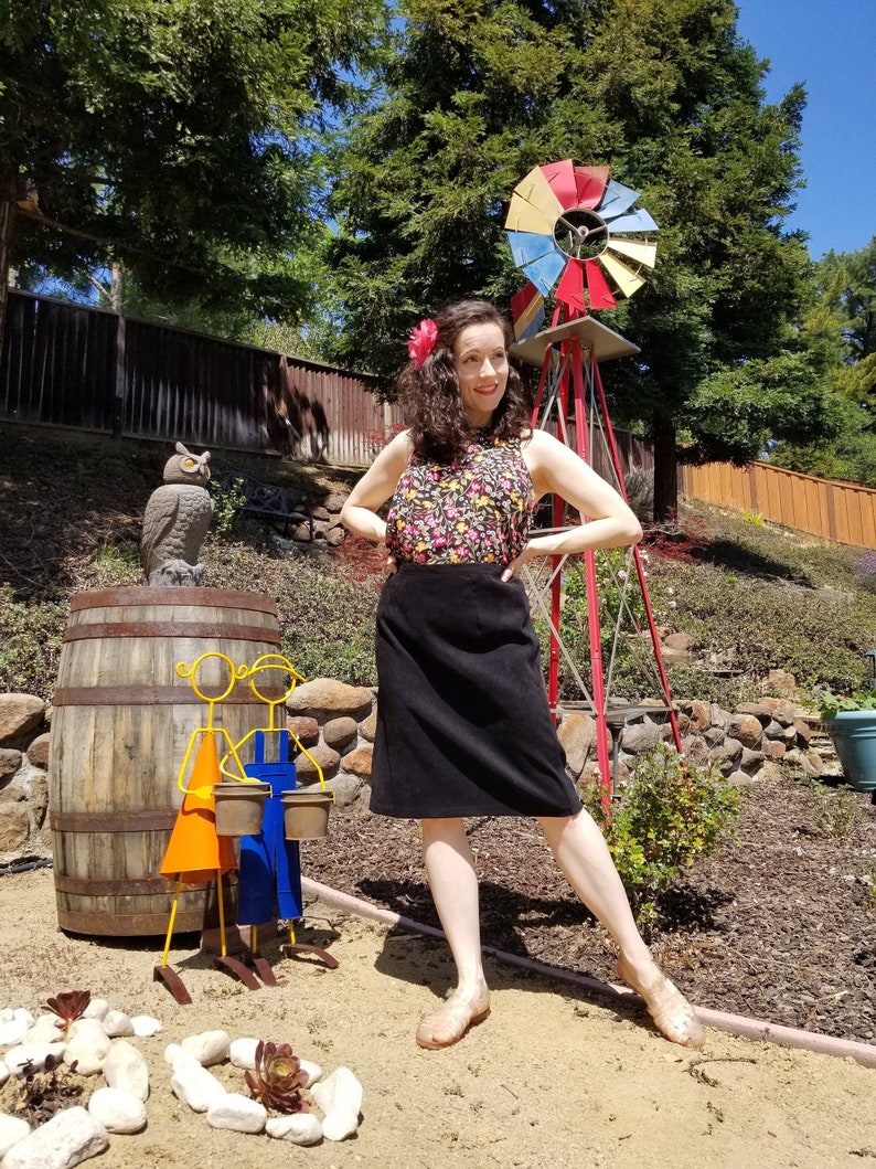 May include: A woman wearing a black skirt and a floral print top stands in a garden setting. She is smiling and looking at the camera. There is a wooden barrel, a windmill, and a small owl statue in the background.