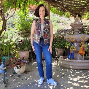 May include: A woman wearing a sleeveless vest with a colorful geometric pattern, blue jeans, and white sneakers. She is standing in a garden setting with a fountain in the background.