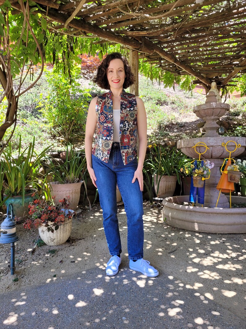 May include: A woman wearing a sleeveless vest with a colorful geometric pattern, blue jeans, and white sneakers. She is standing in a garden setting with a fountain in the background.